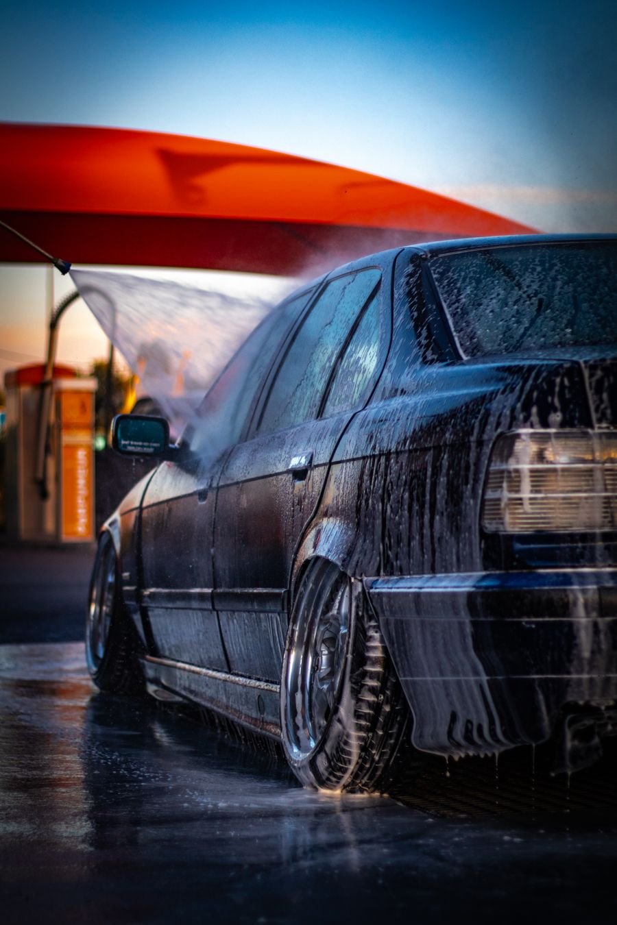 Dark saloon car being rinsed under a wash canopy at dusk